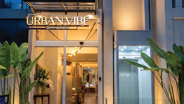 A hotel entrance with a sign reading 'URBAN VIBE BANGKOK,' a glass door, and indoor plants.
