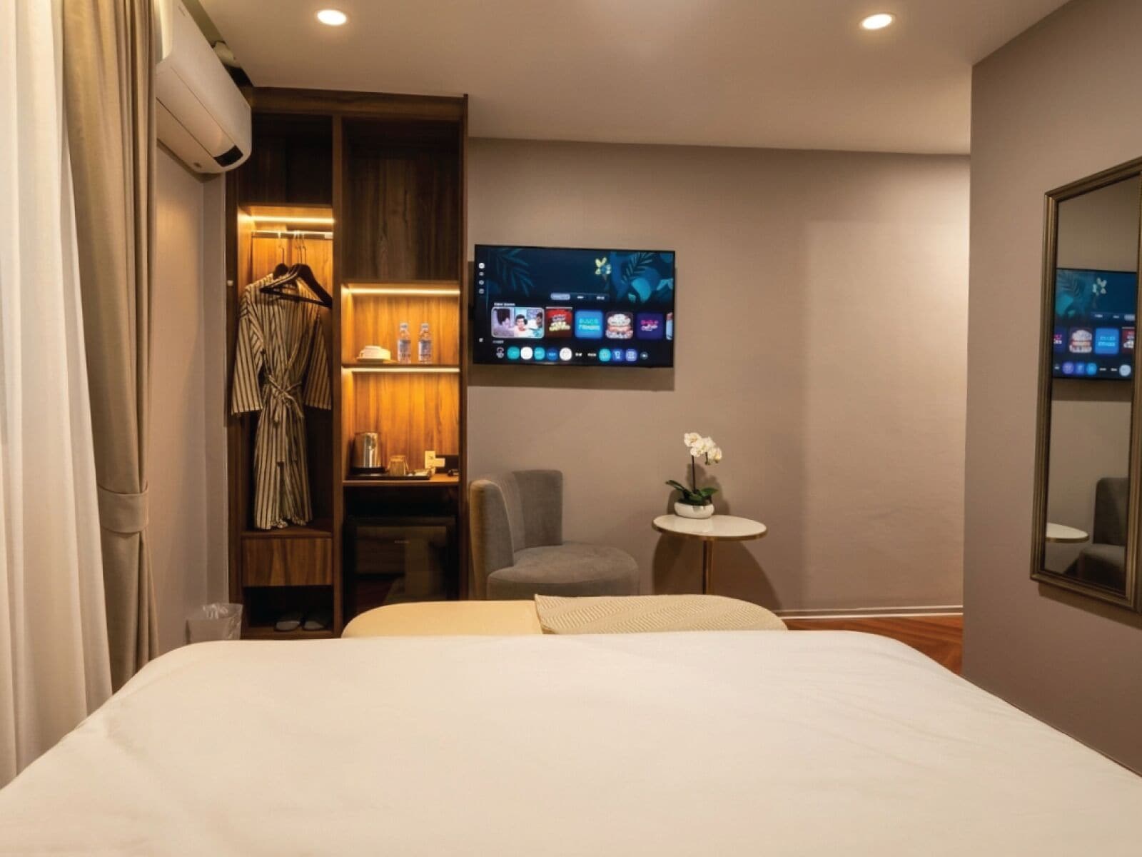 Een hotelkamer met een bed, een aan de muur bevestigde televisie, een tafeltje met een bloem en een kledingkast met daarin hangende kleding.