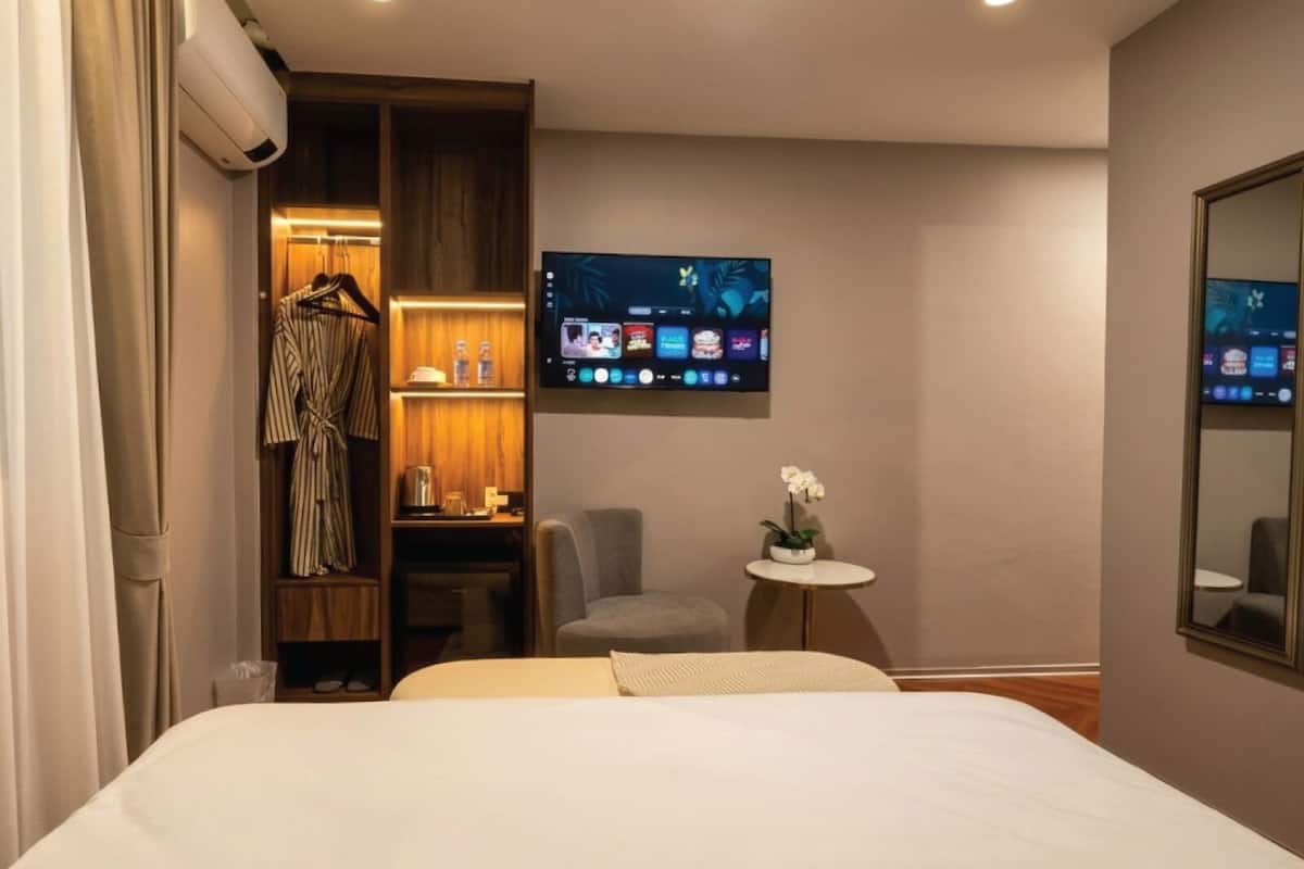 Ein Hotelzimmer mit einem Bett, einem an der Wand befestigten Fernseher, einem kleinen Tisch mit einer Blume und einem Kleiderschrank mit darin befindlicher Kleidung.