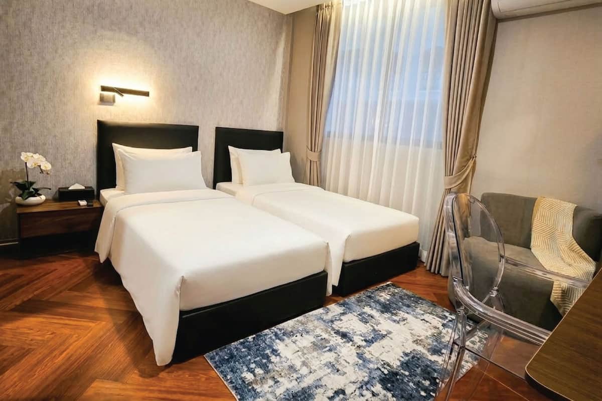 Ein Hotelzimmer mit zwei Betten, einem Nachttisch mit Lampe, einem Sessel und einem großen Fenster mit Vorhängen.