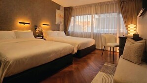 A hotel room with two beds, a desk with a lamp, a chair, and a large window with curtains.