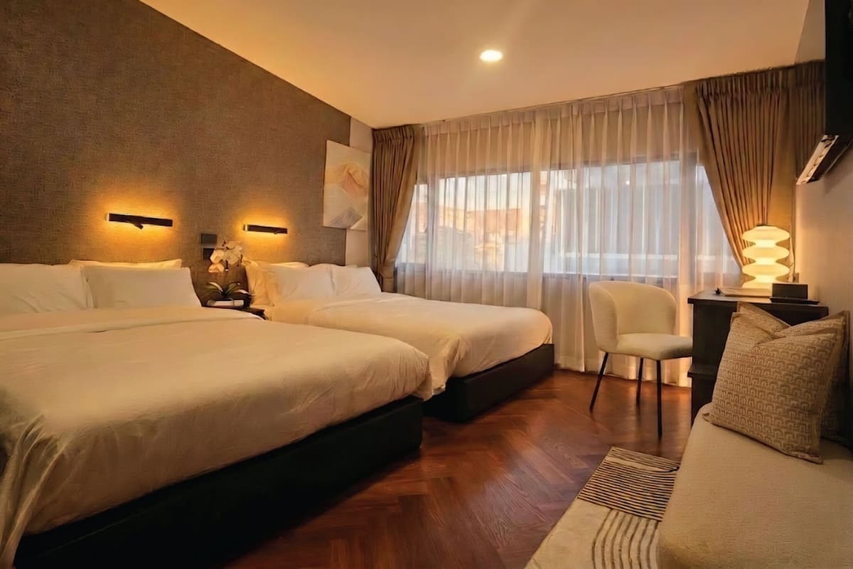 Ein Hotelzimmer mit zwei Betten, einem Schreibtisch mit Lampe, einem Sessel und einem großen Fenster mit Vorhängen.