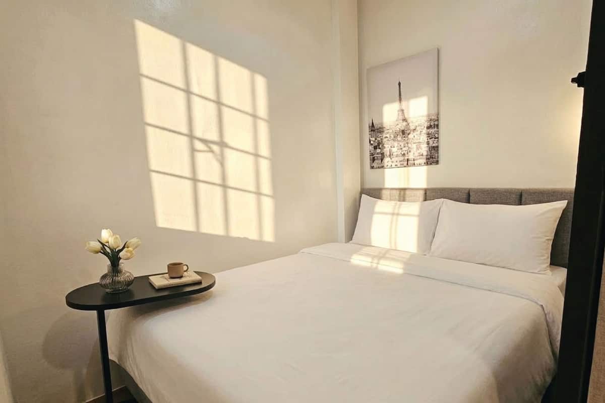Ein ordentlich bezogenes Bett mit weißen Leinen, ein kleiner Nachttisch mit Blumen in einer Vase und einer Tasse, sowie ein gerahmtes Stadtbild an der Wand.