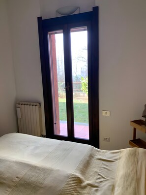 A room with a bed, a radiator, a door with a view to the outside, and a small wooden side table with a lamp. - Holiday Home 'Vacanze' with Lake View, Wi-Fi and Air Conditioning (Laveno-Mombello)