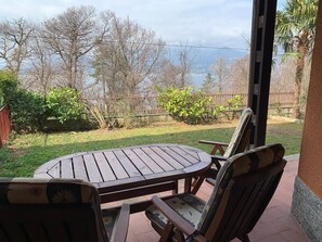 Eine Terrasse mit rundem Tisch und Stühlen, Blick auf Bäume und ein Gewässer, sowie eine Zäunung.