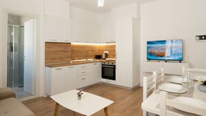 Une cuisine moderne avec des armoires blanches, un comptoir en bois et un four encastré. On y trouve un coin repas avec une table et des chaises blanches, ainsi qu’un téléviseur à écran plat fixé au mur.