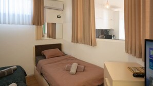 Habitación de hotel con una cama individual, un escritorio con computadora y control remoto, una ventana con cortinas, un equipo de aire acondicionado y un espejo.