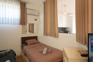 Hotelkamer met een eenpersoonsbed, een bureau met een computer en afstandsbediening, een raam met gordijnen, een airconditioner en een spiegel.