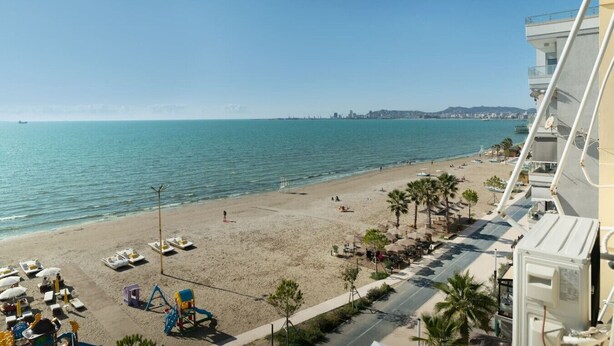 A beach view from a hotel balcony with a sandy beach, palm trees, and a clear blue sea. - Greenview Apartment by Tok Apartments (Durrës)