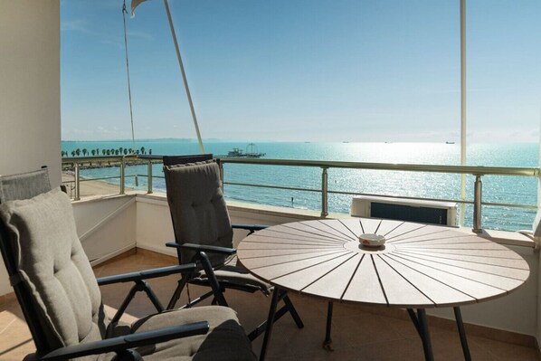 Een balkon met een ronde tafel en twee stoelen, met uitzicht op de kust, de zee en een schip in de verte.