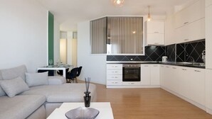 A modern kitchen with white cabinets, a black backsplash, and a built-in oven. There is a dining area with a white table and chairs, and a gray sofa in the living room. - Greenview Apartment by Tok Apartments (Durrës)