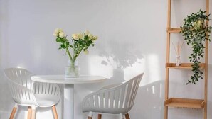 Ein Zimmer mit einem runden weißen Tisch, zwei weißen Stühlen mit Holzbeinen, einer Vase mit weißen Blumen und einem Holzregal mit Grünpflanzen.