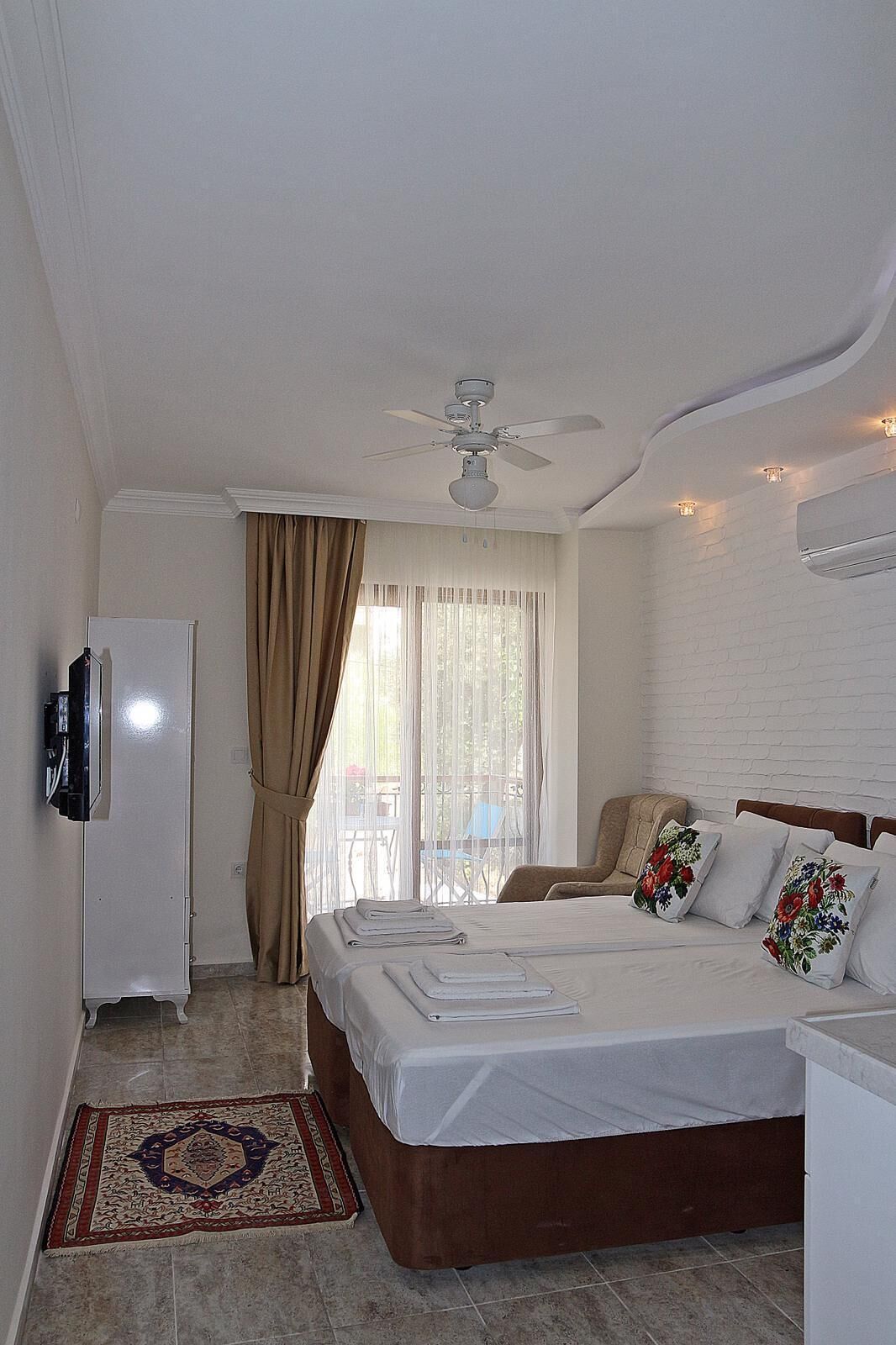Ein Hotelzimmer mit Bett, Deckenventilator, Fernseher, Fenster mit Vorhängen und einem kleinen Tisch mit Stuhl.