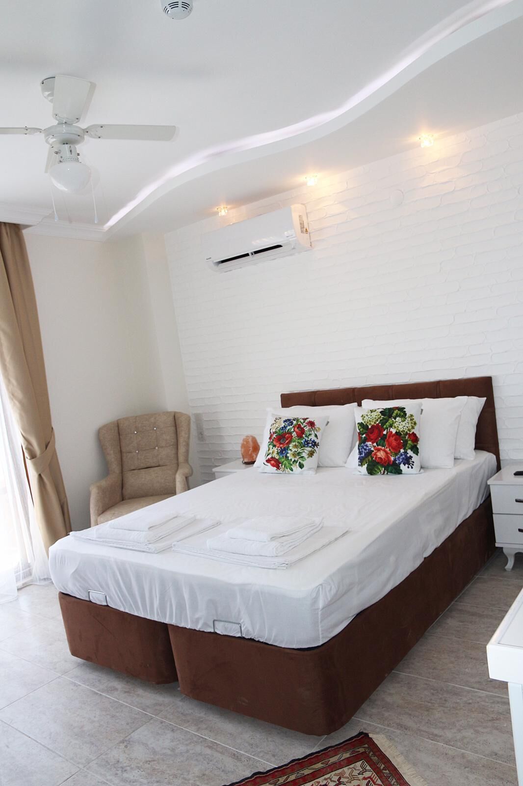 A neatly made bed with white linens, a brown headboard, and floral pillows. There is a beige armchair, a bedside table with a salt lamp, and a white nightstand. The room has white brick walls, a ceiling fan, and an air conditioning unit.
