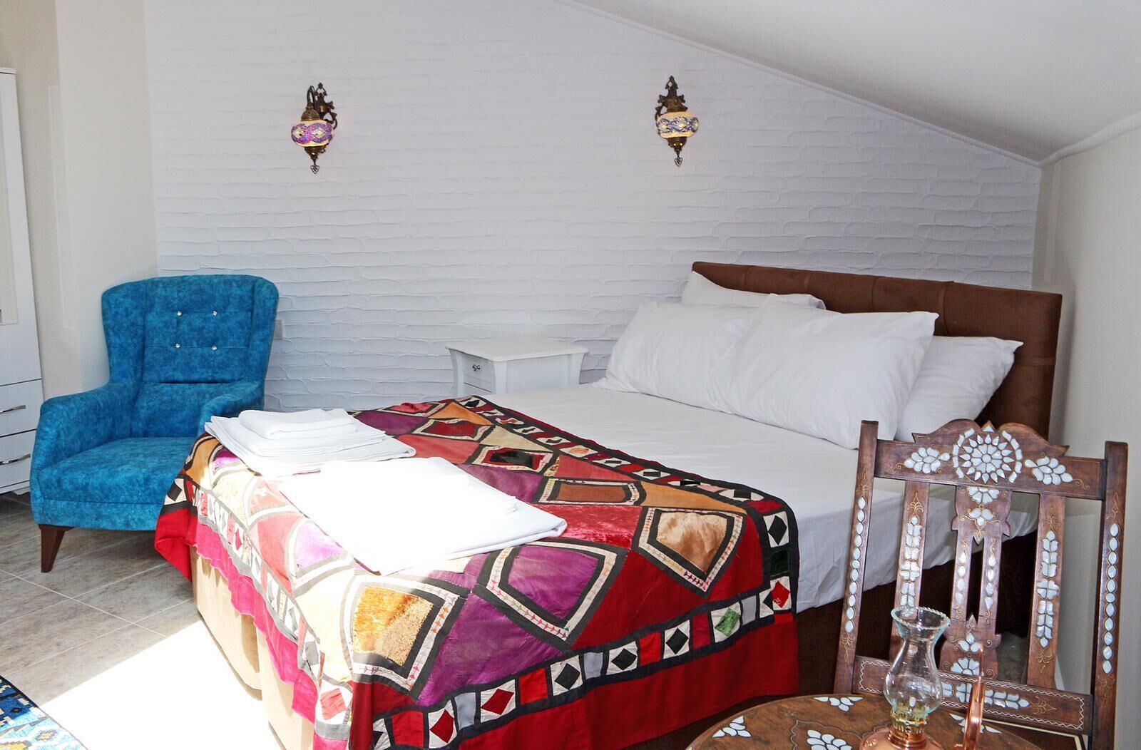 A bedroom with a bed, a blue chair, a small table, and decorative wall sconces.