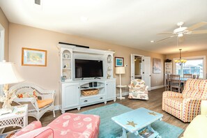 A living room with a television, white wicker furniture, a patterned sofa, and a dining area with chairs. - Beachside Bungalow - Dog Friendly - Amazing views from rooftop deck! (Topsail Beach)