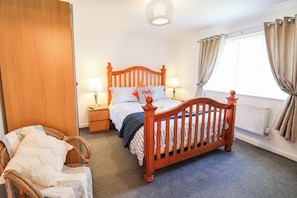 A bedroom with a wooden bed, two bedside lamps, a wicker chair, and a window with curtains. - Seaview (Lowestoft)
