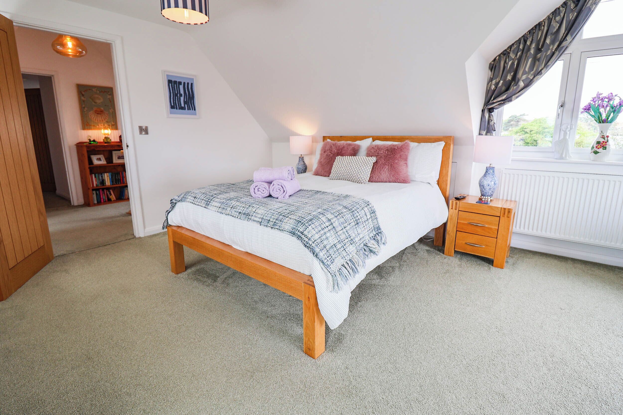 A bedroom with a wooden bed, a nightstand, a lamp, a window with curtains, and a bookshelf.
