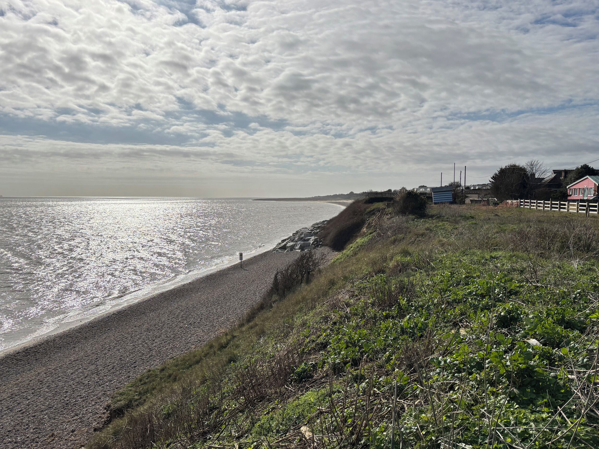 Eine Küstenlandschaft mit einem kiesigen Strand, einer grasbewachsenen Böschung und einem entfernten Blick auf ein Haus mit rotem Dach.