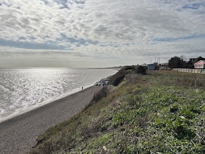 A coastal landscape with a pebbly beach, a grassy embankment, and a distant view of a house with a red roof. - Seaview (Lowestoft)