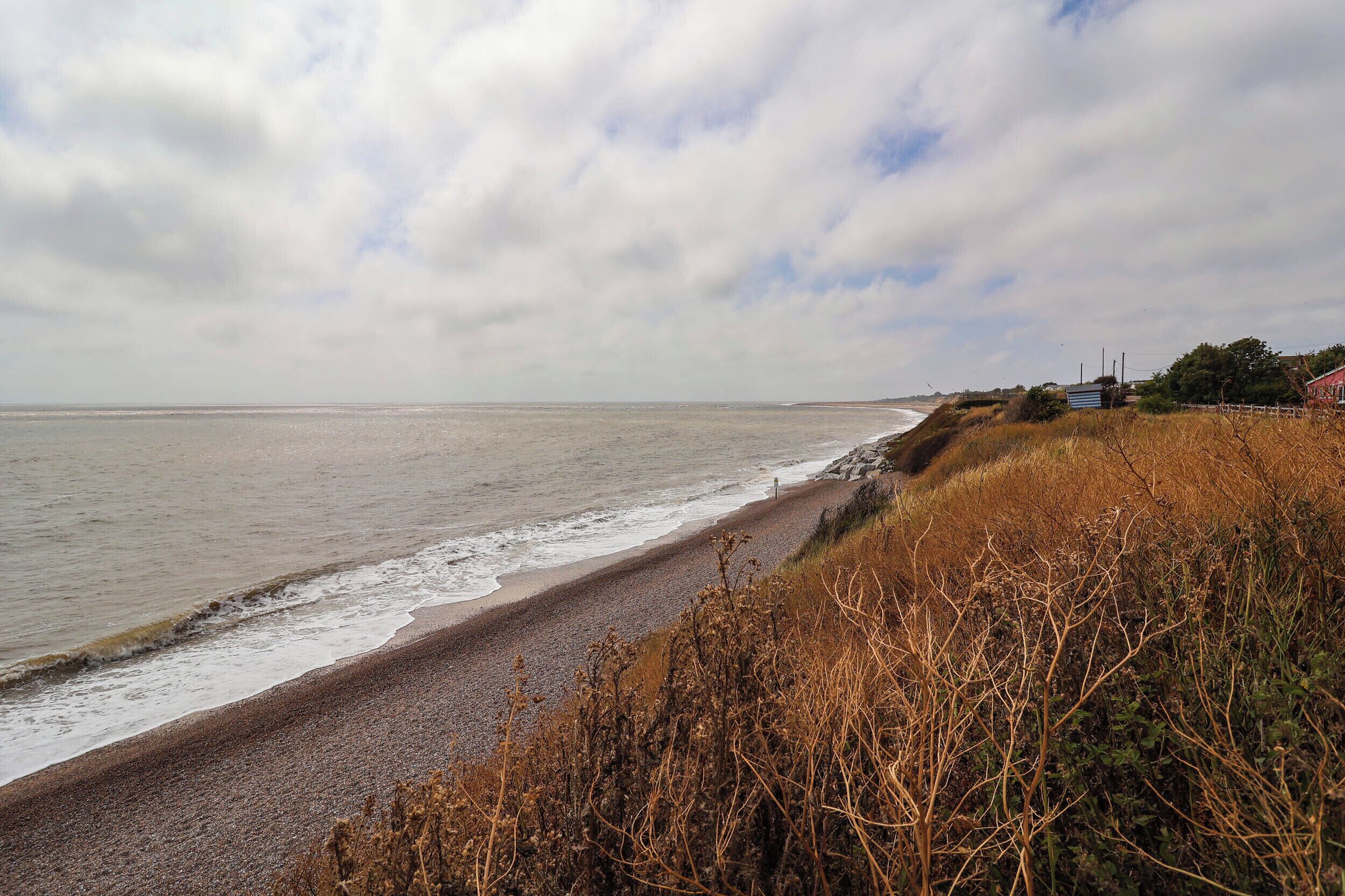 A coastal landscape with a pebbly beach, overcast sky, and sparse vegetation.