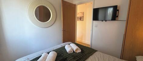 Una cama bien hecha con toallas enrolladas, un espejo redondo, un televisor en la pared y una puerta que da a otra habitación.