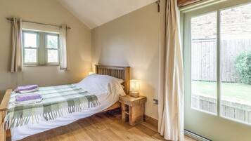 Ein Schlafzimmer mit einem Holzbett, einem Nachttisch mit Lampe, einem Fenster mit Vorhängen und Blick auf einen Holzzaun sowie Grünfläche draußen.