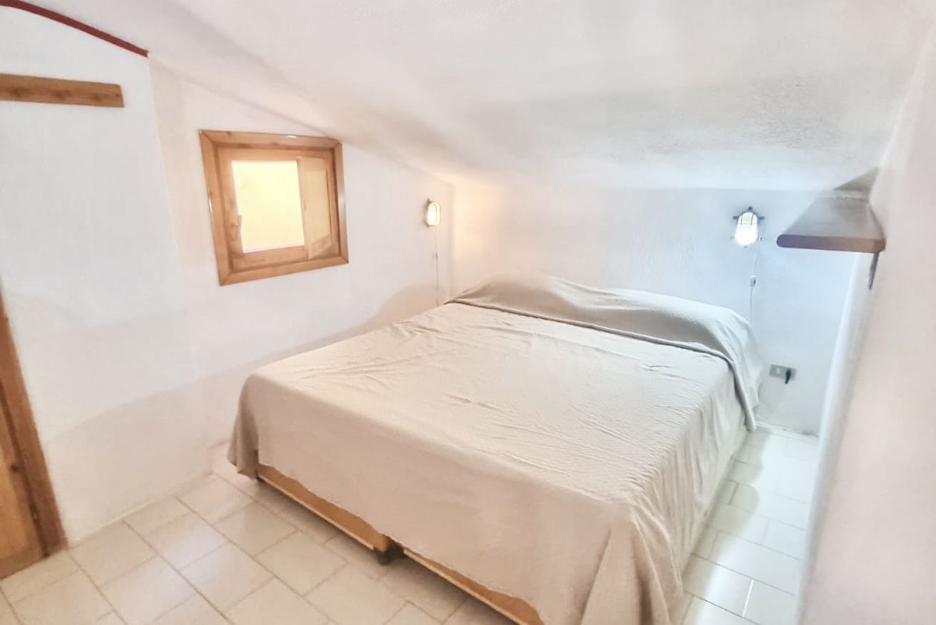 Una camera angusta con un letto singolo, una testiera in legno, una piccola finestra e un punto luce fissato alla parete.