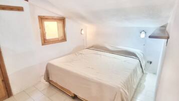 Una camera angusta con un letto singolo, una testiera in legno, una piccola finestra e un punto luce fissato alla parete.