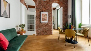A room with a brick wall, a green sofa, a dining table with chairs, and a window with curtains. - BDC - NoLo Industrial Loft 2Bath (Milan)