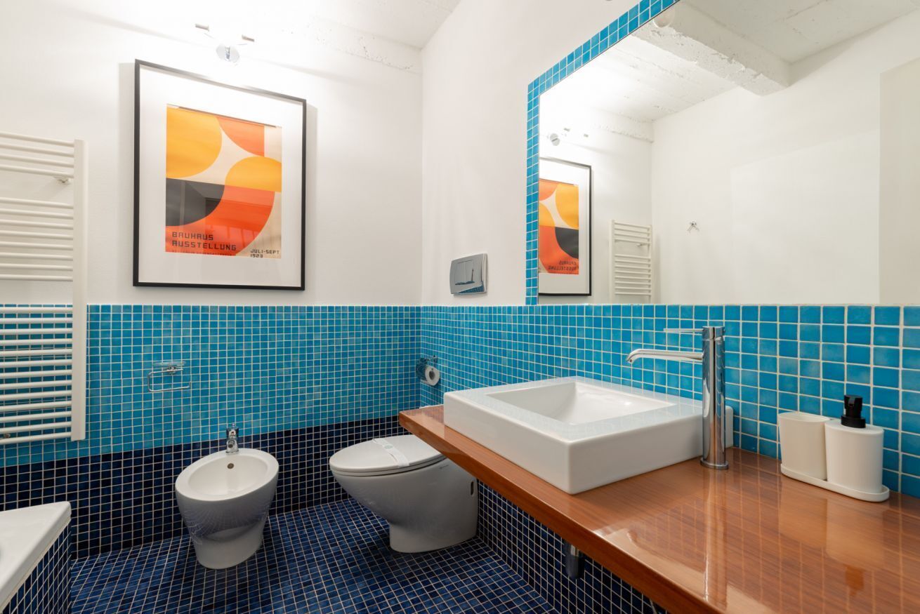 Ein Badezimmer mit einem weißen Waschbecken, einem Bidet und einer Toilette. An den Wänden befinden sich Bilder in Rahmen, und über dem Waschbecken hängt ein Spiegel.