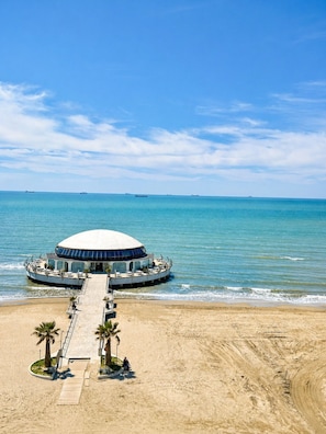 A beachfront building with a dome structure, a pier leading to it, and a sandy beach with palm trees.