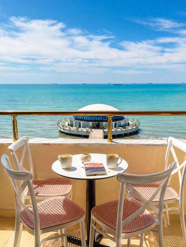 A balcony with two white chairs and a small table, overlooking a calm sea and a circular building with a dome.
