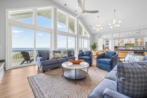 A spacious living room with a large window overlooking the ocean, a ceiling fan, and a modern kitchen in the background. - Stargazer 10 bedroom Brand New Luxury Home on Hatteras Island (Hatteras)