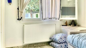Ein Schlafzimmer mit Fenster, Heizkörper, einem Bett mit gemusterten Kissen, einem Nachttisch und einer Hängelampe.