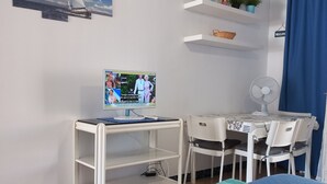 Une chambre d’hôtel équipée d’un lit, d’un bureau avec un ordinateur, d’une chaise, d’un ventilateur et d’un téléviseur diffusant une émission.