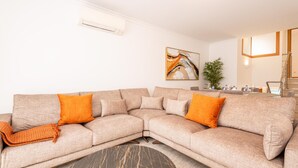 En moderne stue med en beige hjørnesofa, to orange pynsepuder, et sofabord i sort marmor og et maleri på væggen.