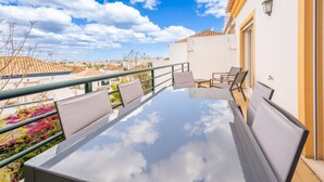 En balkon med et glasbord og stole, der ud til et boligområde med huse og en klar himmel.