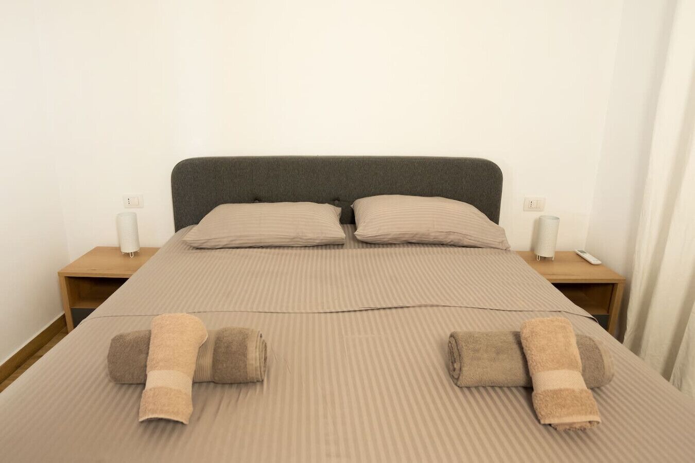 A neatly made bed with striped bedding, two pillows, and two rolled towels. There are two bedside tables with lamps and a remote control on one.