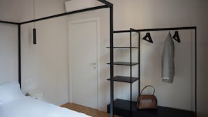 Une chambre avec un lit, une porte blanche, une étagère en métal noir contenant des vêtements et un sac à main, et une petite poubelle blanche.