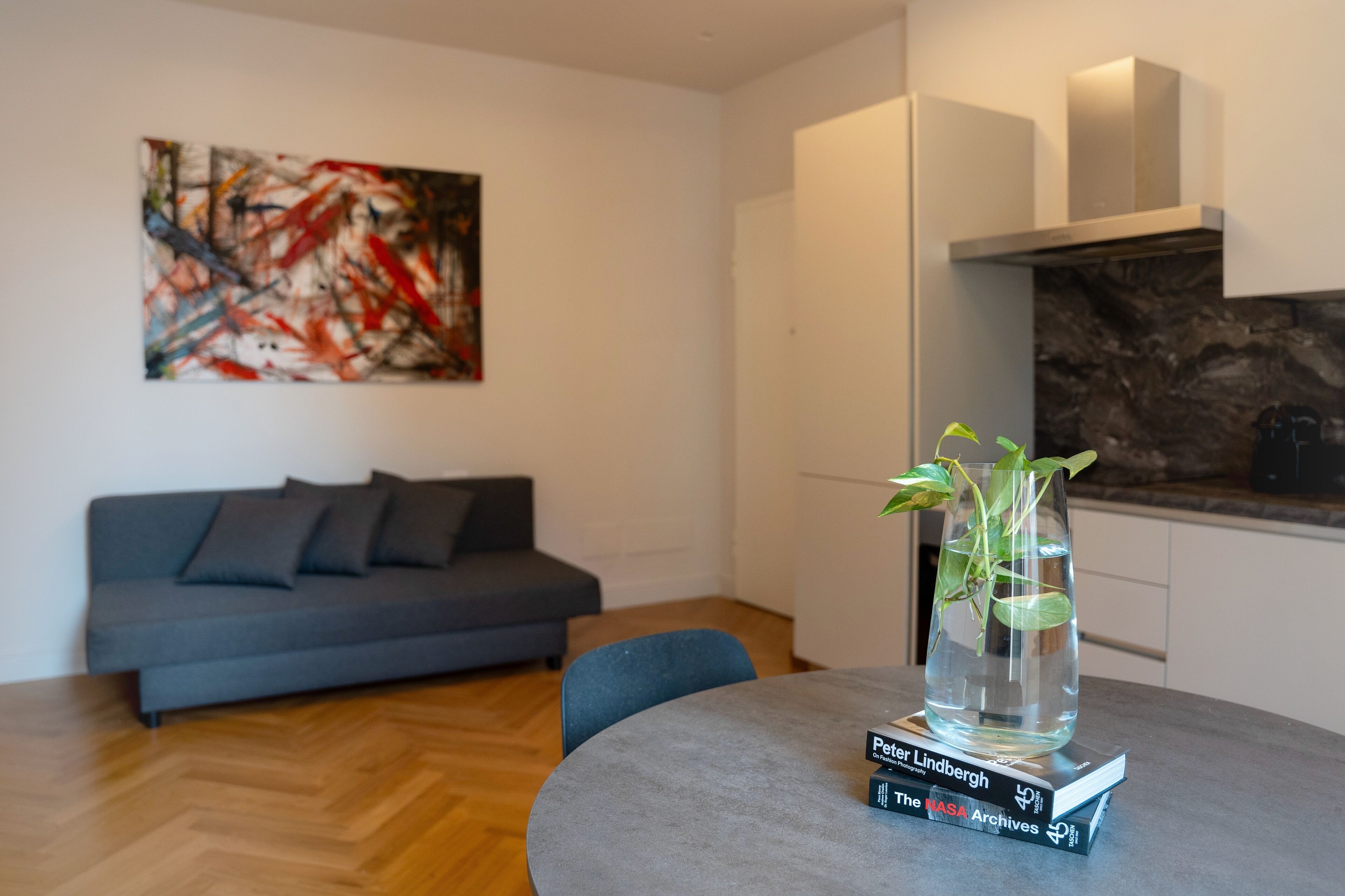 Een moderne woonkamer met een grijze bank, een ronde tafel met een vaas en boeken, en een keuken met een achterwand van steen.