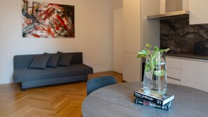 Un salon moderne avec un canapé gris, une table ronde agrémentée d’un vase et de livres, et un coin cuisine doté d’un pare-feu en pierre.