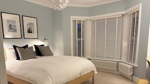 Une chambre avec un grand lit, du linge de lit blanc, une tête de lit en bois, deux tableaux encadrés au mur et une fenêtre avec des volets blancs.