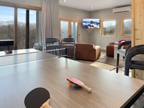 Ein Zimmer mit Tischtennisplatte, Stühlen, einer Couch und einem Fernseher, der einen Blick auf Berge zeigt.