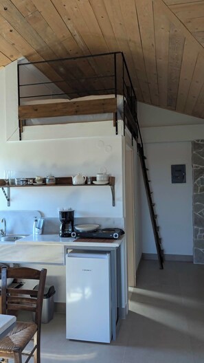 Eine kleine Küche mit integriertem Kühlschrank, einer Kaffeemaschine und einem Herd. Das Zimmer hat eine Holzbalkendecke und eine Steinwand.