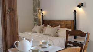 Een keurig opgemaakt bed met kussens, een nachtkastje met theepot en kopjes, en een stoel.