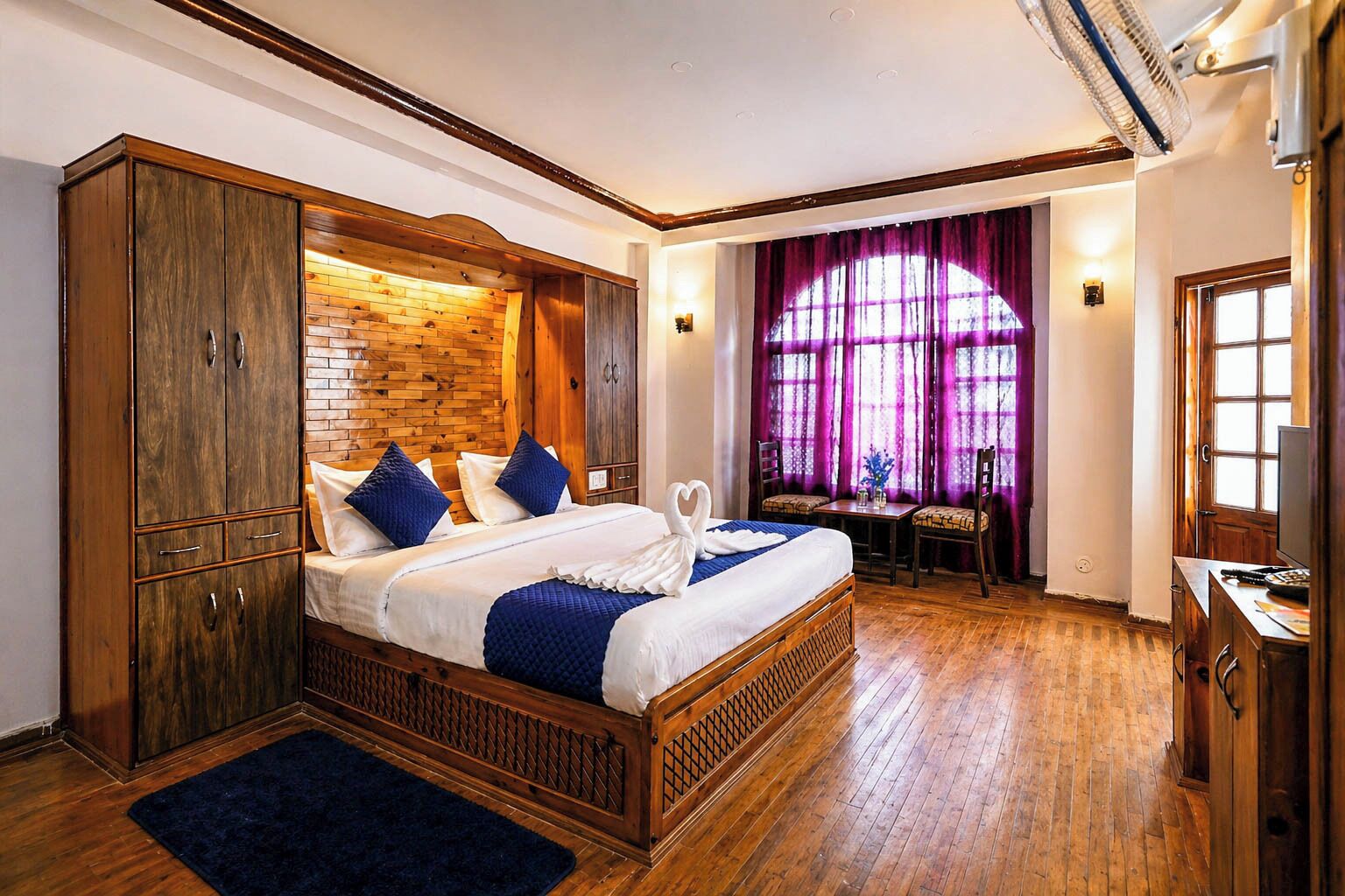 A double bed with a wooden headboard, a small table with chairs, and a window with purple curtains.