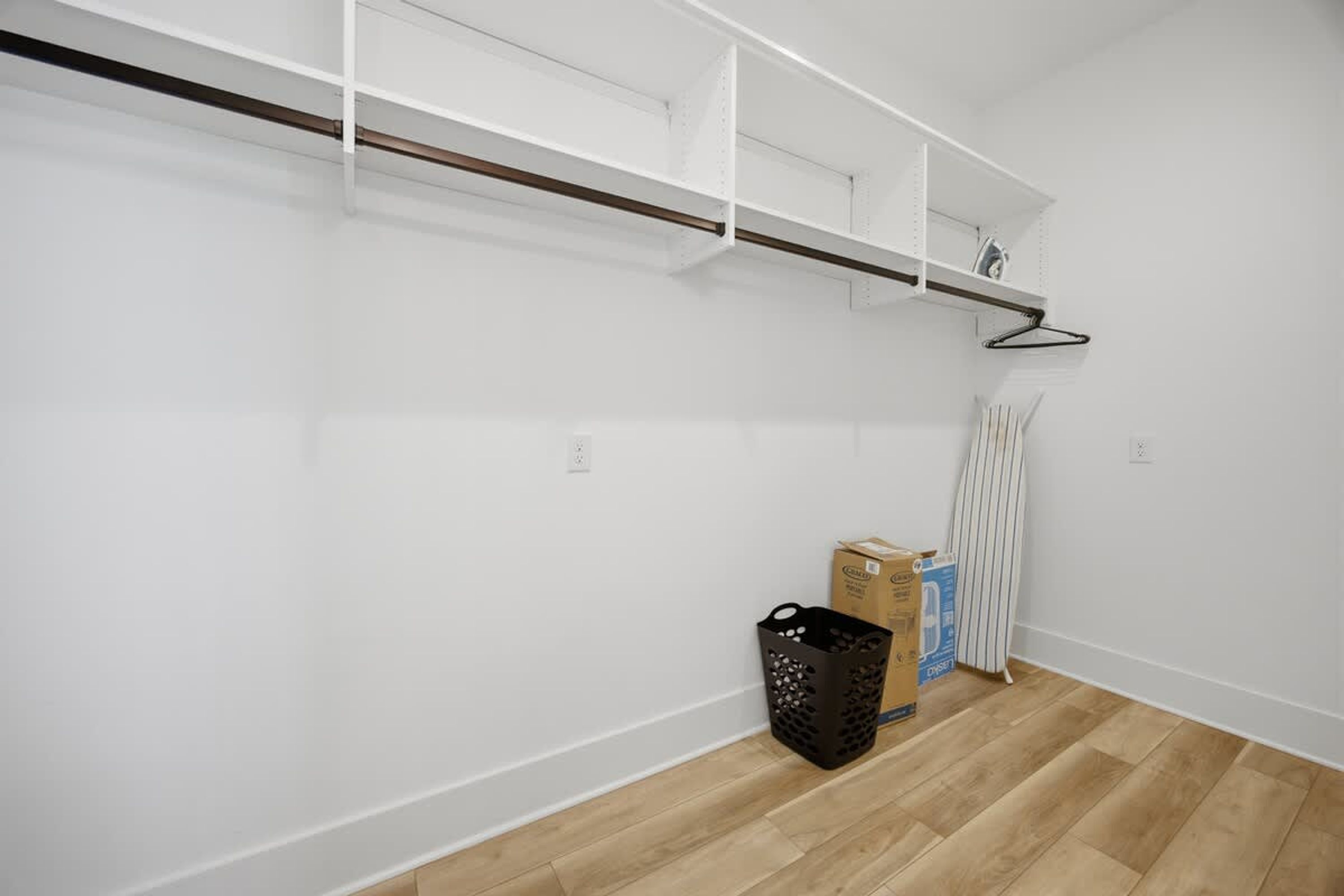 Ein Zimmer mit weißen Wänden, Holzboden und weißen Regalelementen. Auf dem Boden stehen zwei Kartons und ein schwarzer Korb.