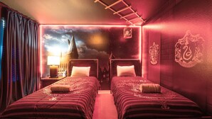 Una habitación con dos camas, una mesita de noche con lámpara, un mural en la pared y una escalera.