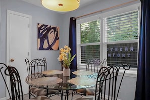 A dining area with a round glass table, six metal chairs, a vase with flowers, a painting on the wall, and a window with blinds. - Charming Townhouse Retreat w/ Patio & Private Balcony Escape (North Charleston)